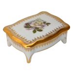 Pequena caixa para joias de porcelana branca com aplicações em dourado à volta e flores ao centro da tampa, com pés. No fundo as inscrições Meissne – Limoges, France 