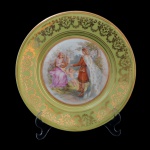 Prato de porcelana tcheca, Manufatura Puls, com borda verde e decoração dourada. Cenas românticas ao centro e borda do círculo interno pontilhada em dourado em alto relevo. Início do século XX. 24,5 cm.