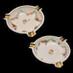 Par de cinzeiros de porcelana da Bavária. Fundo de superfície branca com decoração de guirlandas de flores. No fundo as inscrições: Schumann, Bavária. 11 cm.