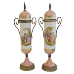 Par de vasos altos, em porcelana europeia, com bojos cilíndricos ornamentados em policromia com cenas românticas. Alças laterais em volutas. Bases e tampas de cor rosa com adornos dourados. Séc. XX. 29 cm.