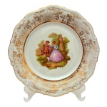 Prato em porcelana Bavária em fundo branco com detalhes em dourado e representação de cena galante de ilustração de Fragonard, borda recortada. No fundo a inscrição Bavária e a letra `F`. 26 cm.
