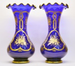BACCARAT - Par de vasos em cristal de tom azul cobalto. Apresenta decoração monocromática ouro, exibindo largas faixas entrelaçadas, envolvendo arranjos florais. Borda soprada em ondulados e finalizada por filetados. França. Séc. XIX. 30 cm.