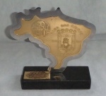Placa comemorativa dos 50 anos das casas da beira em metal prateado sobreposto com metal dourado, formato do mapa do Brasil sobre base de granito preto. Alt. 15cm