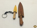 Lote de 2 miniaturas de facas em aço com pega em madeira e bainha em couro. Medindo a maior 14cm de comprimento.