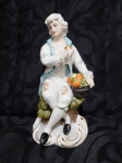 Figura de porcelana representando vendedor de frutas com aproximadamente 25 cm de altura e em bom estado de conservação.