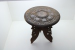 Mesa oriental em madeira fartamente entalhada, tampo circular, pernas curvas e articuláveis. Decoradas com flores, folhas e frutos e ao centro marchetado em bronze. Med. 39x37 cm.