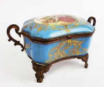 SEVRES  LINDA CAIXA EM PORCELANA COM GUARNIÇÕES EM BRONZE. TAMPA TEM RESERVA COM CENA IDÍLICA CASAL COM GAIOLA DE PÁSSAROS E SEU CÃO. ASSINADA LYSEL.PROFUSAMENTE REMATADO EM OURO. FUNDO NA TONALIDADE AZUL CELESTE. MARCAS DA MANUFATURA SOB A BASE. FRANÇA, SEC. XIX. 23 CM DE COMPRIMENTO.