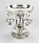 PRATA DE LEI GEORGE V -  SUNTUOSA EPERGNE EM PRATA DE LEI COM MARCAS PARA CIDADE DE SHEFFIED E LETRA DATA 1911. PRATEIROS WALKER E HALL ESTABELECIDOS EM 1894. ESTILO E ÉPOCA ART DECO. CONSTRUÍDA POR UM PRATO CENTRAL ELEVADO SOBRETRÊS ALTOS PÉS. DO PRATO CENTRAL PENDEM TRÊS OUTROS PRATOS DE MENOR TAMANHO. INGLATERRA, 1911. 30 CM DE ALTURA. 1730 GNOTA: NOTA: A palavra epergne é tirada do francês epargne, que significa economia, e originalmente exibiu a aparência de ortografia mais anglicizada. A epergne inglesa economiza de duas maneiras. Primeiro, na economia de espaço precioso: o costume de serviço no sec. XVIII e inicio do XIX requeria que grande parte dos alimentos fossem colocados na mesa ao mesmo tempo. Os comensais que entram na sala de jantar encontraram a comida no lugar, geralmente em pratos de entrada cobertos (os pratos, às vezes, eram colocados sobre um recheou térmico com água quente), e era composto por um ou dois tipos de peixe e uma ou duas sopas. Além disso, por volta de 1760, tornou-se moda para o anfitrião esculpir o pássaro ou outro animal que fosse a opção de carne na mesa (lembra-se dos filmes em que as mesas tinham porcos inteiros, pássaros com penas para enfeitar montados no centro?).A segunda maneira em que economiza a epergne é que ela torna econômico o uso de nozes raras, frutas, condimentos e outros luxos dos trópicos. Os convidados se serviam da epergne, e as iguarias que não eram comidas ficavam intocadas nas mesas centrais, em vez de serem desperdiçadas quando os pratos fossem limpos no serviço convencional. Assim as EPERGNES podem conter frutos, doces, petiscos e outras iguarias para serem consumidas com os dedos e também se prestam para decoração com flores. A partir do período vitoriano a epergne foi aliviada de sua grande posição de servidor.No "novo" estilo de serviço, a la russe, cada prato era trazido separadamente em sequencia, deixando mais espaço para a exibição de flores neste acessório. O final do século XIX e início do século XX viu um breve e limitado ressurgimento de epergnes nos estilos rococó e neoclássico, ainda assim, estes foram destinados a serem usados como objetos decorativos, não propriamente para servir.