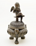 Caixa em bronze fundido e cinzelado, encimada pela figura de querubim alado, repousando sobre recipiente esférico, ornamentado por rostos angelicais contrapostos entre raiados. Base tripoide de representação zoomorfa. Séc. XIX. 20 cm.