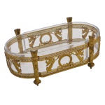 Centro de mesa em metal amarelo dourado de formato retangular e cantos arredondados, guarnecido de recipiente em fino cristal translúcido. Suspenso por quatro colunas em formato de aljava para flechas. Encontra-se apoiado em quatro piões. Nos dois frisos fenestrados que percorrem a borda e a base, estão fixados quatro pares de figuras aladas, que ladeiam coroa de louros. Séc. XX. 6 x 24,5 x 13 cm.