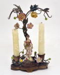 Candelabro de manufatura italiana concebido em metal forjado em representação de árvore, guarnecido de apliques de flores e figura de camponesa em porcelana policromada. Base recortada apoiando duas bobeches contrapostas ao centro. Itália. Séc. XX. 25 x 15 cm.