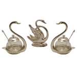 Conjunto de três peças em metal, sendo: A) Par de porta geleias na forma de cisnes acompanhadas de suas colheres da Casa Chase Importação. (16 x 12 cm); B) Porta guardanapo também na forma de cisne.  (15 x 13 cm). Novos. Sem uso. Na caixa. Pertenceu ao Espólio de Vitória Dib Bachour.