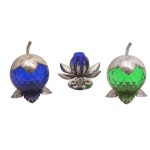 Conjunto de três peças em metal prateado com recipientes de vidro, sendo:  Par de potes para geleia na forma de frutos nas cores verde e azul (12 x 8 cm) e 1 castiçal na forma de flor se abrindo, na cor azul. (8 x 8 cm). Bases e tampa de metal. Pertenceu ao Espólio de Vitória Dib Bachour.