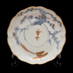 Prato de porcelana provavelmente europeia, apresentando decoração com borda onduladas e friso dourado. Reserva central com pássaro em dourado e arvoredo e folhagens na cor azul. No fundo selo da coleção: Nobrega Antiguidades. 21 cm.