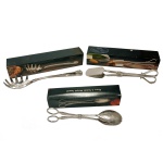 Conjunto de três peças de metal prateado, sendo: A) Duas pinças para salada (25 cm e 20 cm). B) Pegador de macarrão (28 cm). Novos, sem uso. Caixa original. Pertenceu ao Espólio de Vitória Dib Bachour.