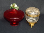 Duas peças : a) Pequeno baleiro em vidro italiano de Murano na tonalidade rubi, pega da tampa em flores e folhagens(com restauro). Etiqueta de fabricação no fundo. Alt 13 x diam 12 cm. b) Pote em vidro translúcido e acidado, tampa esmaltada com elementos Art Nouveau, guarnições de metal amarelo. Alt. 10 x diam. 8cm.