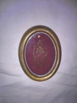 Quadro oval em resina dourada, com decoração de estrelas e volutas vazadas em metal dourado, sobre feltro na cor vinho e sob vidro, medindo 13 x 10 cm.