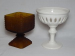 Duas fruteiras: a) Em cerâmica branca, haste na forma de torneado, galerias vazadas. Pequeno bicado na borda. 17 x 15cm. b) Em vidro fosco âmbar, ornamentado com desenhos em alto relevo de cachos de uvas. Rachado na parte interna. 14 x 10 x 10cm.
