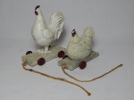Galo e galinha em resina branca repousando sobre carrinho com barbante para puxar. Falta rodinhas traseiras da galinha. 15 x 13 x 5cm e 10 x 12 x 5cm.