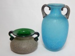 Dois vasos em vidro: a) Vaso estilo ânfora em azul, fundo azul claro fosco, decoração escamosa, alças em preto. Apresenta desgastes. Alt. 20cm. b) Vaso baixo em vidro verde, decoração externa em chapisco, alças em volutas. Alt. 9cm.