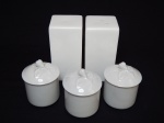 Cinco peças em porcelana branca: a) Saleiro e pimenteiro, formato retangular. 10 x 5 x 5cm. b) Três potinhos arredondados, tampa com pega no formato de folhas e frutas. 7 x 6cm.