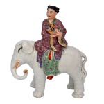 Estatueta de porcelana japonesa, policromada, representando menina com cesta de flores sentada em elefante. 30 x 23 cm.