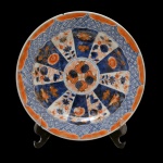 Prato de porcelana japonesa com decoração Imari. Borda lisa decorada na cor rouge de fer e centro com intensa decoração de flores e fruto. Verso com superfície branca com três reservas florais. No fundo selo da coleção: Nobrega Antiguidades. ... 23 cm.  Apresenta bicado.