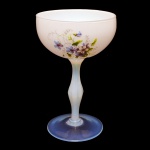 Taça de opalina francesa na cor branca leitosa, decorada com elementos florais em tons de lilás, roxo e verde. Haste em losango e base translúcida. 22 x 14 cm.