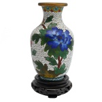 Vaso de procedência chinesa em metal esmaltado, em cloisonné, trazendo desenhos de arbustos floridos, sobre fundo branco ilustrado. Borda dourada, com base de madeira na cor preto cerejeira. 13 cm / 15 cm com base.
