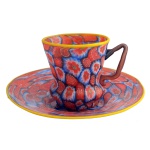 Xícara para café em formato peculiar, acinturado, pires de amplo diâmetro, com estampas assimétricas, em tons predominantes de vermelho e azul escuro. Borda amarela em ambas as peças; alça em tom acobreado com pinceladas em vermelho e azul, formando conjunto pouco convencional, porém homogêneo. 6 x 7.5 cm / 12 cm pires.