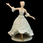 Estatueta de figura feminina em porcelana europeia, em vestido branco esvoaçante, modelo tomara que caia, com detalhes ricamente esculpidos; sapatos e pingente com cordão dourados ao pescoço nu. 24 x 19 cm.