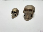 2 esculturas na forma de crânio humano em metal dourado. Medindo a maior 8,5cm de comprimento x 5,5cm de altura.