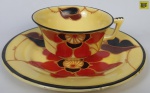 Xícara para chá com prato para bolo em em porcelana art deco, esmaltado com decoração floral, sobre fundo amarelo. Prato apresenta bicado.
