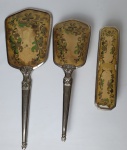 Três peças para toucador em metal prateado, sendo suporte para espelho e duas escovas. Medindo: 25 x 8 cm, 17 x 4 cm e 31 x 11 cm.