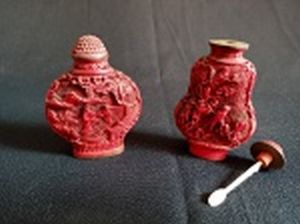 Dois Snuff-Bottle chineses, vermelhos, bojos esculpidos, um com pássaros e folhagens e um com figuras em jardim. Bases de metal com ideogramas.Uma das tampas presa. Alts. 6,5 e 7,5 cm.