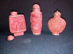 Três Snuff-Bottle chineses, vermelhos, bojos esculpidos com figuras em jardim. Bases de metal com ideogramas. Uma das tampas com a rolha partida e as outras presas. Alts. 6 - 6,5 e 7,5 cm.
