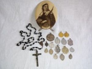 Dezoito peças religiosas diversas: a) Placa oval de metal, estampada com imagem de Santa Luzia. 12,5 x 9,5cm. b) Terço com contas negras e crucifixo de metal. c) Dezesseis medalhas em diversos materiais (1 de prata). Diam. da maior 3,5cm.