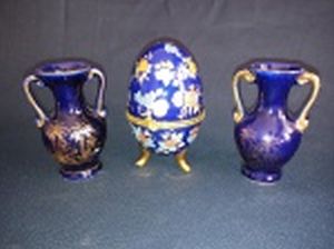 Três peças em porcelana azul cobalto: a) Caixa no formato de ovo de Fabergé, decoração esmaltada com flores, detalhes em douração. Alt. 10cm. b) Par de vasos em miniatura detalhes e decoração de palmeiras filetados a ouro. Alt. 8cm.