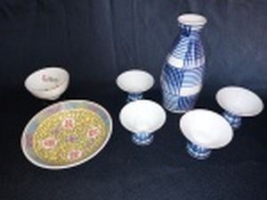 Sete peças em porcelana oriental: a) Conjunto para saquê japonês, decorado cmo faixas azuis sobre fundo branco, composto por garrafa e quatro copinhos, um com bicado. Alt. garrafa 13cm. b) Dois bowls, um com peanha. Maior 3 x 11cm.