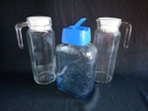 Três peças: a) Duas jarras para suco, vidro translúcido e oitavado, tampas plásticas. Alt. 24cm. b) Garrafa para água, vidro moldado com frutas, tampa plástica. Alt. 23cm.