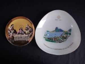 Dois pratinhos decorativos : a) Em porcelana japonesa, borda em faixa dourada, centro aplicado com gravura da A Santa Ceia em transfer-printing. Leves perdas na douração. Diâm. 13cm. b) Em vidro triangular, comemorativo do centenário do Rio de Janeiro (1965), aplicado com desenho da vista da Baía de Guanabara. Diâm. 19cm.