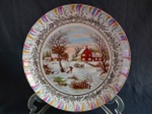 Prato decorativo em porcelana branca, borda e cinturão de folhagens filetados a ouro, centro aplicado com cena rural no inverno. Marca no fundo não identificada. Diâm. 25cm.