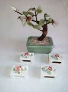 Cinco peças em porcelana: a) Vaso verde Celadon com árvore representando bonsai, folhas de vidro colorido. Algumas perdas. Alt. 16cm. b) Quatro porta caixas de fósforos aplicadas com flores. Alguns quebrados. No estado. 2 x 5 x 3,5cm.