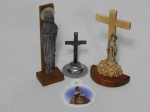 Quatro peças religiosas em diferentes materiais: Cristo cruficiado em metal, 12cm; Maria a frente de cruz em madeira, alt. 18cm; sineta em metal com base de madeira; e placa em porcelana com imagem do Menino Jesus de Praga, 10 x 8cm.