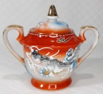 Açucareiro em porcelana oriental de tons branco e `rouge de fér` ricamente decorado por figuras de dragões em alto relevo e filetação dourada. Mede 11 cm de altura por 12,5 cm de uma alça a outra.