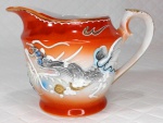 Cremeira em porcelana oriental de tons branco e `rouge de fér` ricamente decorada por figuras de dragões em alto relevo e filetação dourada. Mede 7,5 cm de altura por 11,5 cm da alça ao bico.