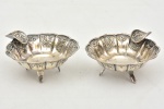 Par de cinzeiros em metal banhado de prata decorado por escudo e figuras de conchas e rocailles em relevo apoiado sobre três pés. Medida: 7,5 cm x 3 cm de altura