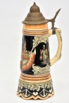 Tankard musical alemão em porcelana policromada, predominando a cor azul cobalto, corpo ornamentado com paisagem montanhosa da cidade de Hamburgo, base recortada circundada por folhas de parreira e cachos e uva em relevo, tampa em metal. Caixa de música na base à corda. Med.: 28 cm x 11 cm