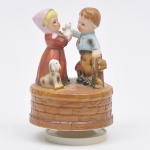 Grupo escultórico em porcelana representando crianças, ricamente policromada, fabricada no Japão, giratória, musical, funcionando.  Medida: 17 cm x 10 cm
