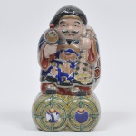Grupo escultórico representado Deus da Fortuna oriental em porcelana esmaltada e ricamente policromada. Medida: 29 cm x 16 cm; Daikokuten era originalmente um deus guerreiro Hindu. Suas primeiras estátuas japonesas eram representadas por uma feição feroz, porém, com o tempo, foram assumindo um rosto risonho. Suas associações à guerra também foram deixadas de lado, tornando-se conhecido como deus da riqueza e da cozinha. Sua virtude é a fortuna.Está sempre em pé ou sobre fardos de arroz e sua protuberante barriga indica que está bem alimentado e próspero. Na mão direita, carrega um martelo de madeira e sobre o ombro esquerdo leva um saco onde carrega tesouros.Há também um mito de que Ebisu é o filho de Daikokuten. Os dois são muitas vezes colocados juntos com um terceiro deus, Fukurokuju, constituindo os Três Deuses da Fortuna.